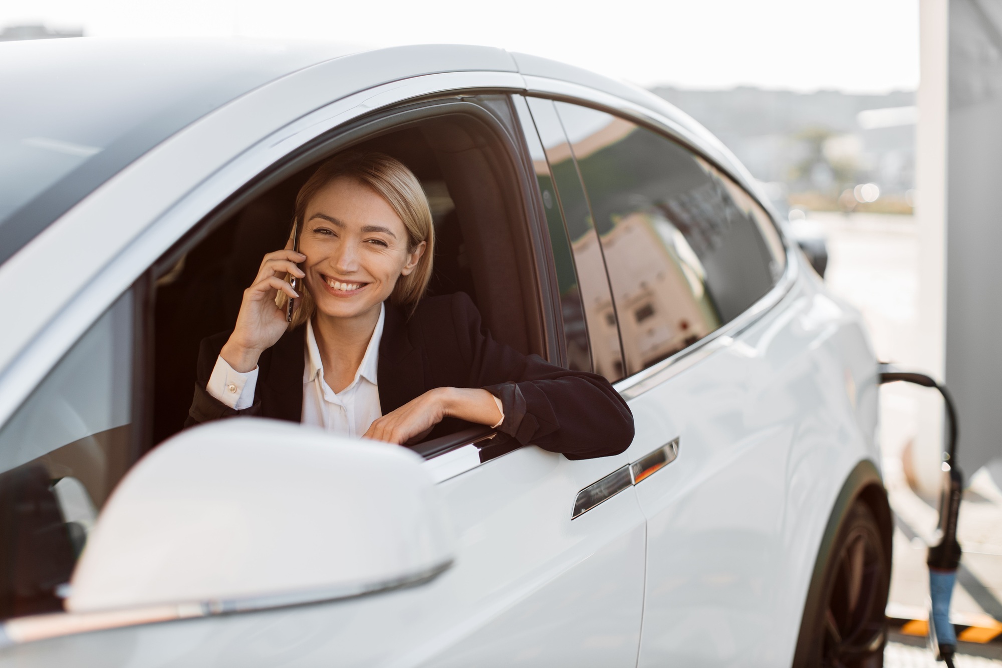 Woman talking on smartphone and charging car on EV station